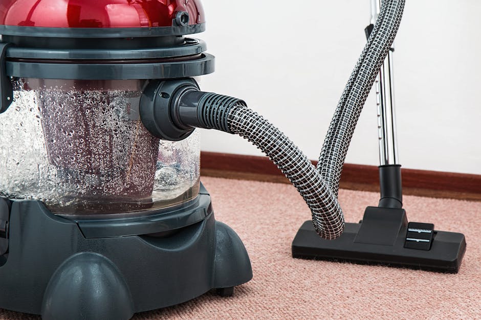 Close-up of a wet vacuum cleaner with a transparent dust container filled with water and debris, connected to a flexible, patterned hose, positioned on a pink carpeted floor in a room with white walls