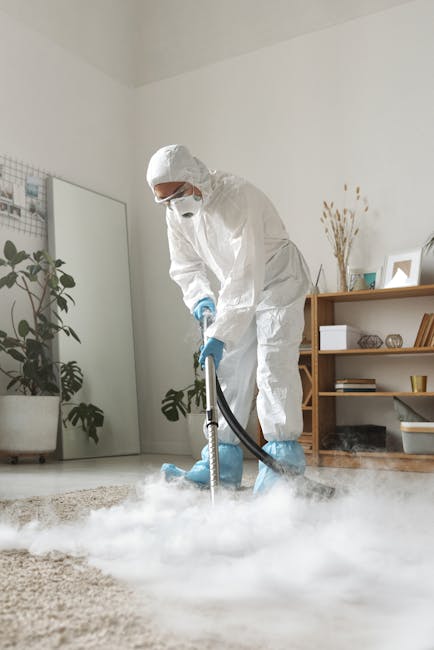 Close-up of a wet vacuum cleaner with a transparent dust container filled with water and debris, connected to a flexible, patterned hose, positioned on a pink carpeted floor in a room with white walls